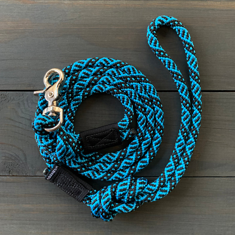 The Teton Quick Clip Leash, a coiled blue and black woven dog leash crafted from durable rock climbing rope with a silver metal clip, is laid out on a wooden surface. The leash features a loop handle and a small black label.