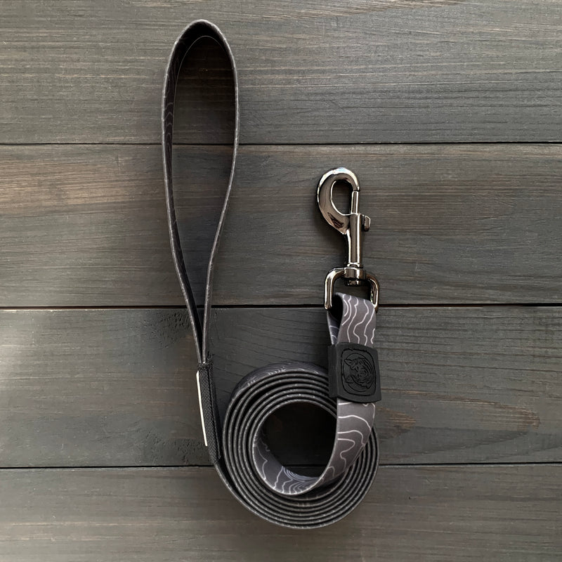 The Black Waterproof Leash + Collar Bundle (x), featuring a stink-proof, wavy-patterned leash with a metal clasp and looped handle, offers durable, everyday dog gear—perfect for walks and shown here on a dark wooden surface.