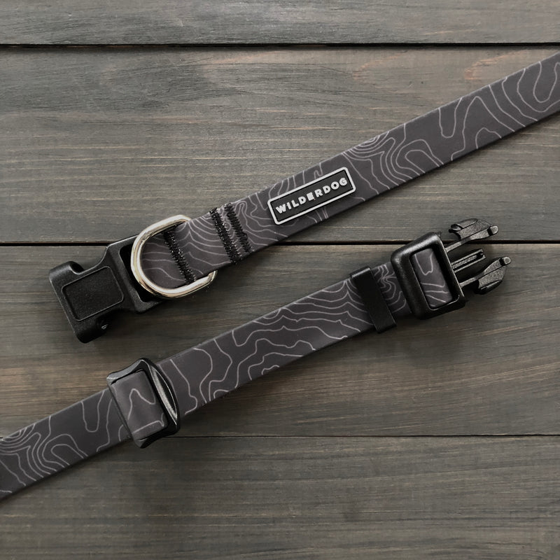 The Black Waterproof Leash + Collar Bundle features durable gear buckles, black topographic lines, a metal D-ring, and a "Wilderdog" label—perfect for any adventure. Two sets are shown resting on a wooden surface.
