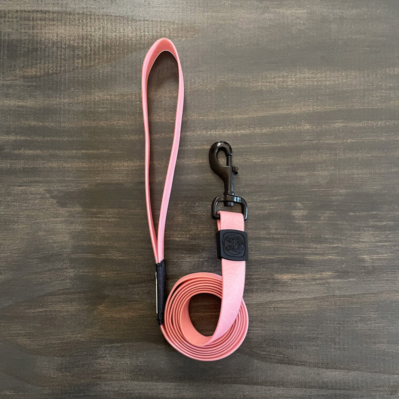 The Guava Waterproof Leash, a durable pink dog leash featuring a loop handle and a black clip on the opposite end, is coiled neatly on a wooden surface. This stink-proof leash also includes a black rubber emblem near the clip.