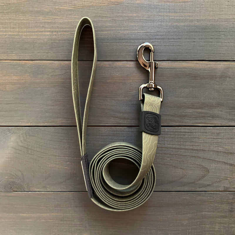 An Olive Waterproof Leash + Collar Bundle (x) with a green patterned, stink-proof leash and silver clip is coiled on a wooden surface. It has a handle loop and black rubber logo, making it essential, durable dog gear.