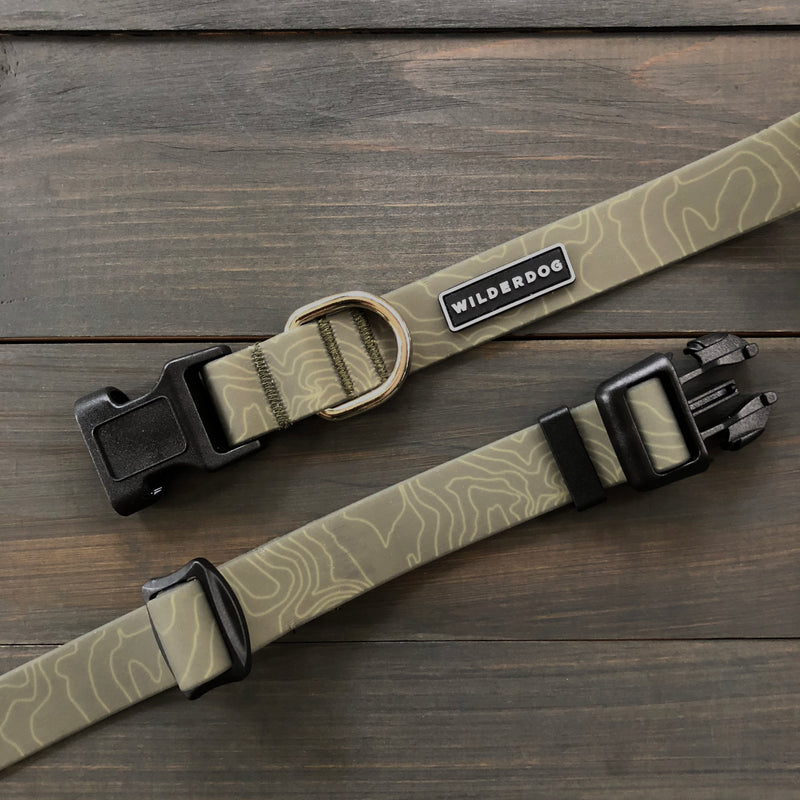 Two Olive Waterproof Leash + Collar Bundles (x) with topographic designs rest on a wooden surface. Each features a black plastic buckle, sturdy metal D-ring, and a label reading "WILDERDOG.