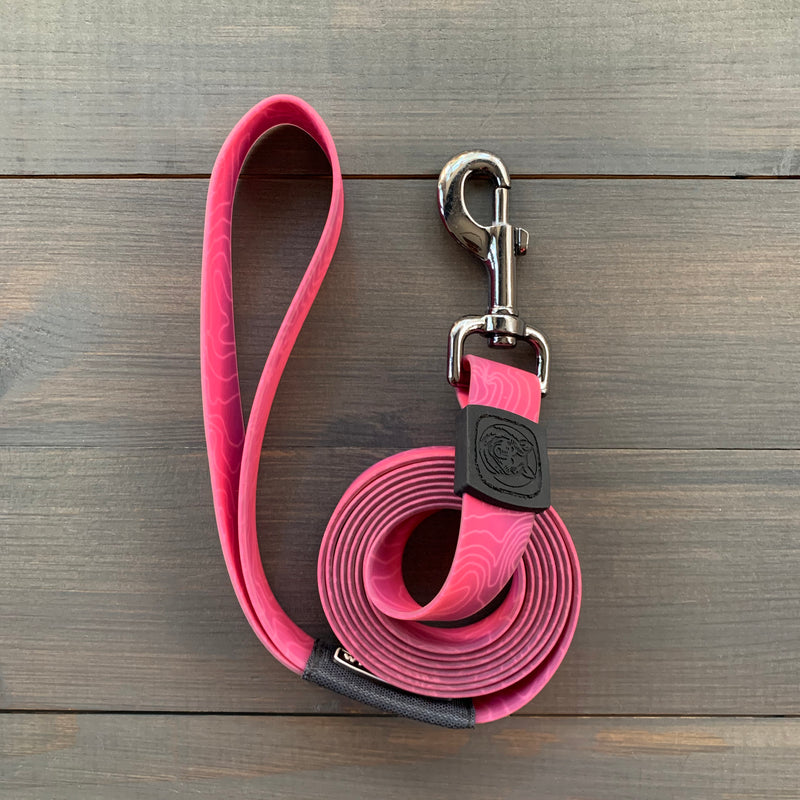 The Plum Waterproof Leash + Collar Bundle (x) is shown coiled on a wooden surface, featuring a pink stink-proof design, looped handle, black rubber logo near the silver clip, and durable construction.