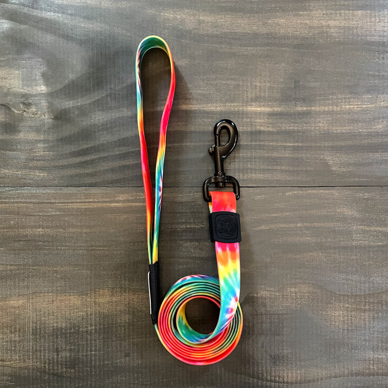 The Tie Dye Waterproof Leash + Collar Bundle (x) features a coiled, stink-proof leash with a vibrant rainbow pattern and black metal clasp, shown on a dark wooden surface—ideal for those seeking durable dog gear.
