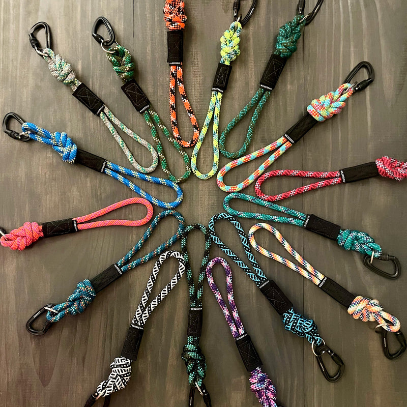 A circular arrangement of Rope Keychains with black locking carabiner clips at one end. Each keychain showcases a unique mix of vibrant colors and designs, radiating from the center of the circle on a dark wooden surface.