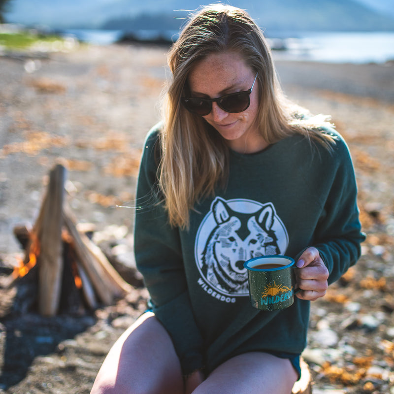 A woman with long hair and sunglasses sits by a small campfire on a beach. She is wearing a green Smartcart21 Wolf Sweatshirt and holding a green mug. The background shows a scenic view of the ocean and distant mountains.